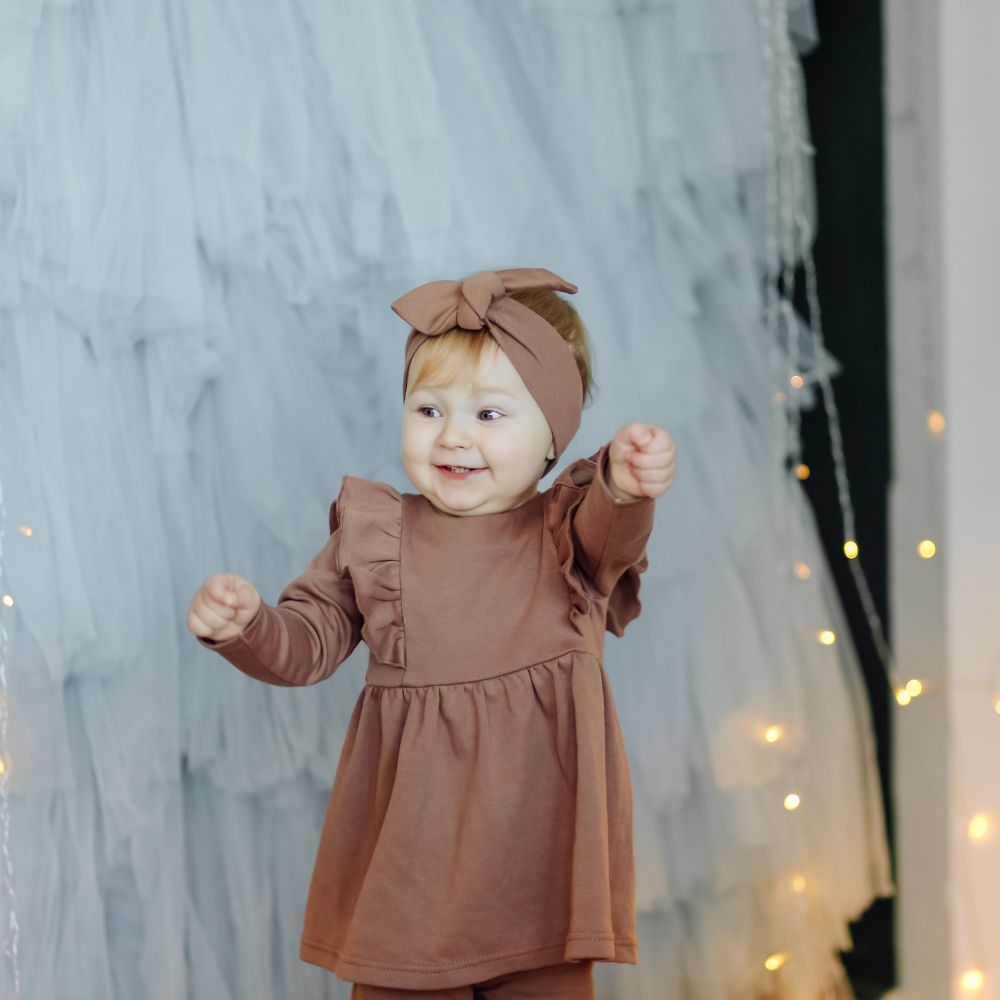 Baby in a brown outfit with a bow headband standing in front of a decorative curtain with lights.