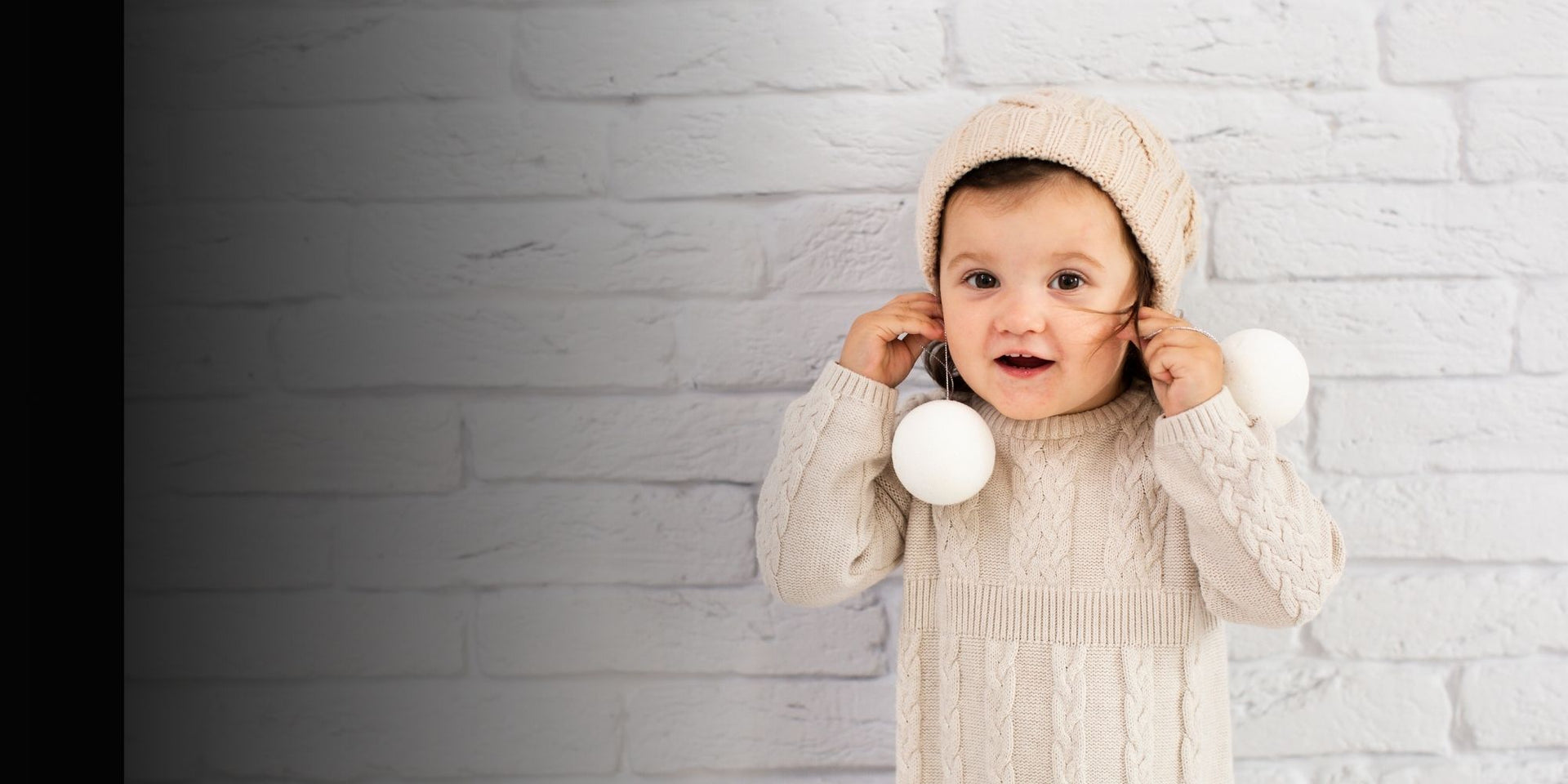 Child wearing a cream-colored knit outfit with white balls against a white brick wall.